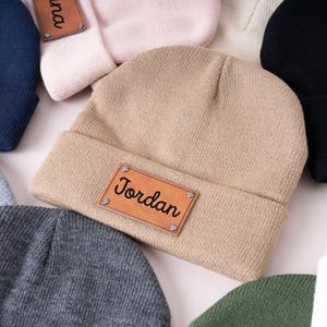 Peut inclure: Plusieurs bonnets en tricot de couleurs variées, dont beige, marine et noir. Chaque bonnet est orné d'un écusson rectangulaire en cuir avec un nom, tel que "Jordan", fixé par des rivets. Les bonnets sont repliés au niveau du bord.
