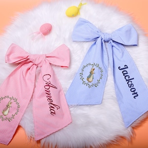 May include: Two gingham fabric bows, one pink and one blue, with personalized names embroidered on the tails. Each bow features an embroidered bunny in a wreath. The bows are on a white fluffy surface with Easter-themed decorations.
