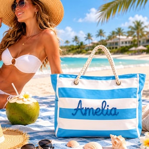 Könnte beinhalten: Eine blau-weiß gestreifte Strandtasche mit dem Namen "Amelia" in blauer Schrift gestickt. Die Tasche hat Seilgriffe und liegt auf einem Strandtuch mit Strohhut, Sonnenbrille und einem Kokosnussgetränk.