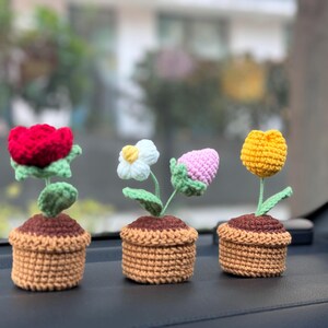 May include: Four crocheted flower pots with different colored flowers. The flowers are red, white, pink, and yellow. The pots are brown and have a crocheted texture.