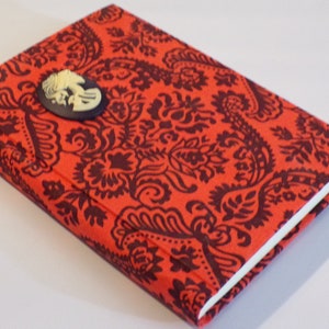 May include: A journal with a red paisley pattern cover and a cameo brooch. The cover is a vibrant red with a dark brown floral design. The cameo is cream-coloured with a dark blue background.