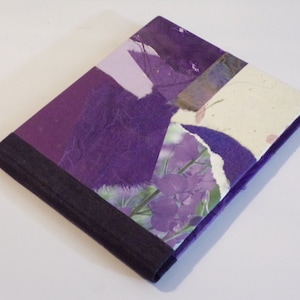 May include: A purple and white handmade journal with a textured cover and a black border. The cover features a collage of paper and fabric with a floral design.