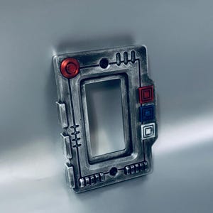 May include: A rectangular, metallic-looking device with a central opening. It features a series of geometric shapes, including a red circle, red square, blue square, and white square. The device has a detailed, industrial design.