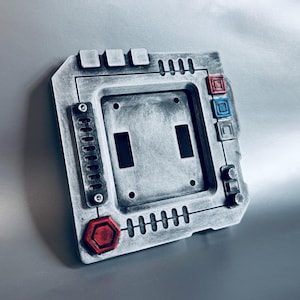 May include: A gray, textured light switch cover with a sci-fi aesthetic. It features two rectangular openings for switches, along with red and blue accents. The cover has a series of small, rectangular and circular details.