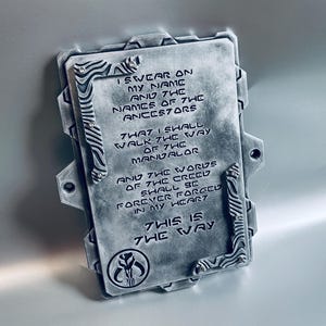 May include: A rectangular, metallic plaque with the text "I SWEAR ON MY NAME AND THE NAMES OF THE ANCESTORS...THIS IS THE WAY." The plaque has decorative corner accents and a Mandalorian symbol.