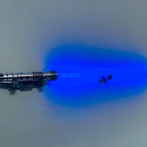 May include: A silver and black toy lightsaber with a bright blue energy blade extending from the hilt. The lightsaber is detailed with various mechanical elements, and the blue light has a slight glow.