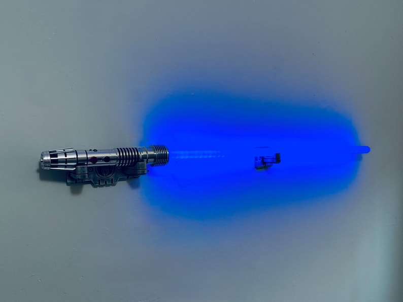 May include: A silver and black toy lightsaber with a bright blue energy blade extending from the hilt. The lightsaber is detailed with various mechanical elements, and the blue light has a slight glow.