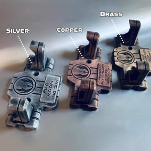 May include: Three rectangular wall mounts in silver, copper, and brass finishes. Each mount features two vertical prongs, a circular Jedi Order emblem, and engraved text reading Plo Koon, Anakin Skywalker, or Luke Skywalker in English and Aurebesh.