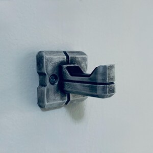 May include: A metallic grey wall hook with a geometric design. The hook has a square base and a curved, open hook extending outwards. The surface has a textured, weathered appearance, suggesting a vintage or industrial style.
