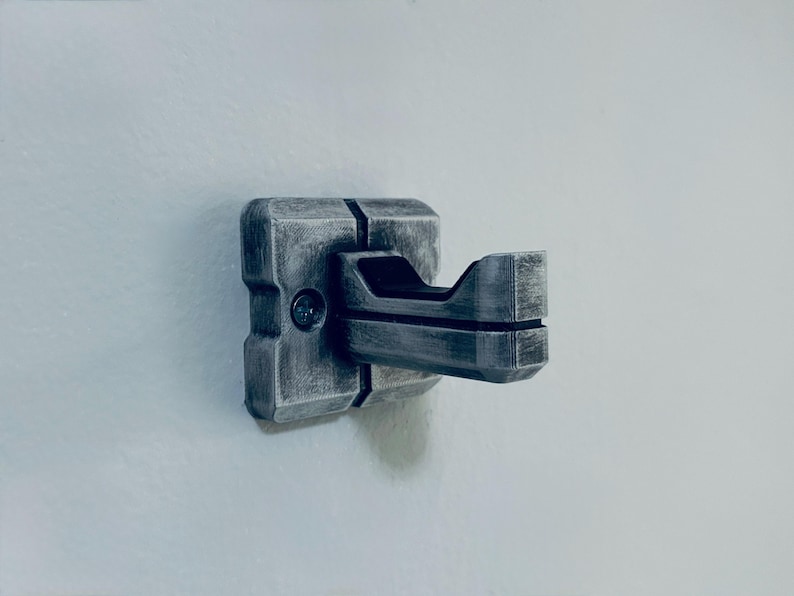 May include: A metallic grey wall hook with a geometric design. The hook has a square base and a curved, open hook extending outwards. The surface has a textured, weathered appearance, suggesting a vintage or industrial style.