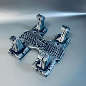 May include: A silver and black weathered display stand featuring four upright supports and a central circular emblem with a starburst design. The H-shaped base has horizontal grooved lines and a metallic finish with dark shading in the crevices.