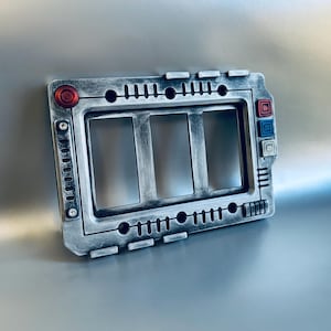 May include: A metallic gray triple light switch cover with a retro-futuristic design. It features three rectangular openings, a red circular button, and blue and red square buttons. The cover has a textured, aged appearance.