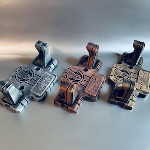 May include: Three rectangular wall mounts with two upward-facing hooks each. The left mount is silver, the centre is copper, and the right is gold. Each features a circular emblem and a rectangular plate with the names Plo Koon, Anakin Skywalker, and Luke Skywalker engraved in English and Aurebesh text.