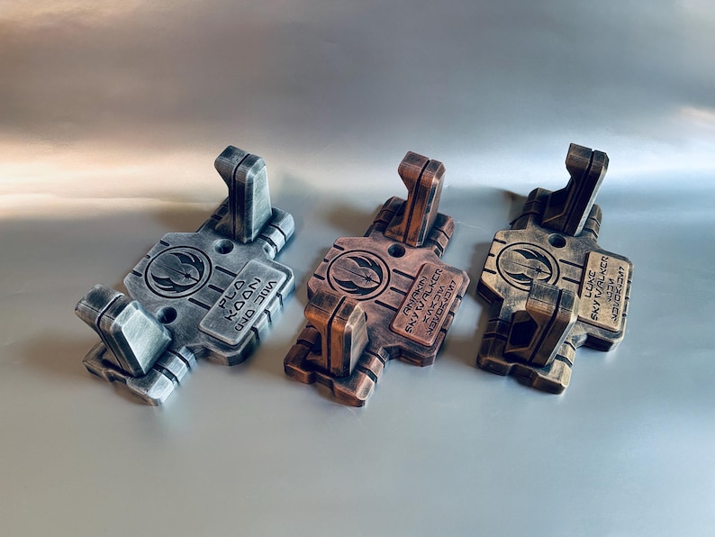 May include: Three rectangular wall mounts with two upward-facing hooks each. The left mount is silver, the centre is copper, and the right is gold. Each features a circular emblem and a rectangular plate with the names Plo Koon, Anakin Skywalker, and Luke Skywalker engraved in English and Aurebesh text.