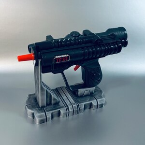 May include: A black Star Wars Jedi blaster toy with a red tip and red trigger, mounted on a gray stand. The blaster has the Star Wars Jedi logo on the side. The stand is gray and has a rectangular base.