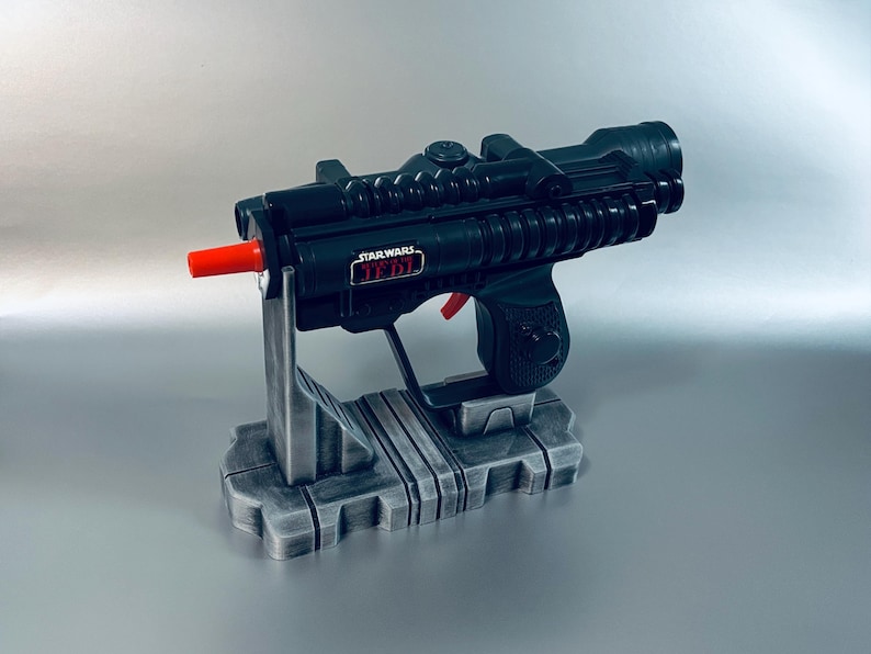 May include: A black Star Wars Jedi blaster toy with a red tip and red trigger, mounted on a gray stand. The blaster has the Star Wars Jedi logo on the side. The stand is gray and has a rectangular base.