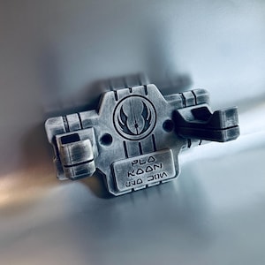 May include: A silver-toned wall mount featuring a weathered metallic finish, designed with two protruding prongs. The centre displays a circular Jedi Order emblem above a rectangular plaque engraved with the text PLA KOON in English and Aurebesh.