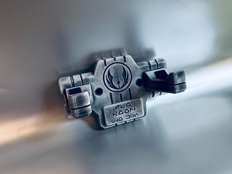 May include: A silver-toned wall mount featuring a weathered metallic finish, designed with two protruding prongs. The centre displays a circular Jedi Order emblem above a rectangular plaque engraved with the text PLA KOON in English and Aurebesh.