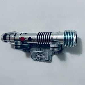 May include: A silver-toned lightsaber hilt with black grip rings, a red activation button, and a black circular accent. The hilt is mounted on a weathered grey display bracket featuring the embossed text "PLO KOON" in a stylized font.