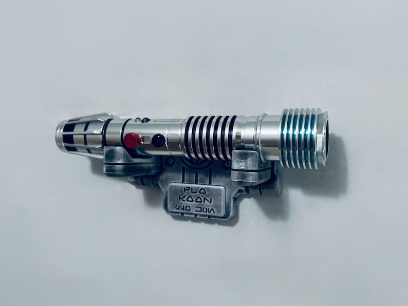May include: A silver-toned lightsaber hilt with black grip rings, a red activation button, and a black circular accent. The hilt is mounted on a weathered grey display bracket featuring the embossed text "PLO KOON" in a stylized font.