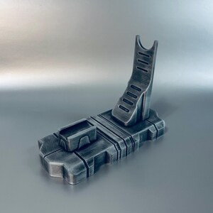 May include: A metallic gray stand with a rectangular base and an angled support. The stand features a small rectangular compartment and a curved, slotted support for holding an item. The design has a futuristic, industrial aesthetic.