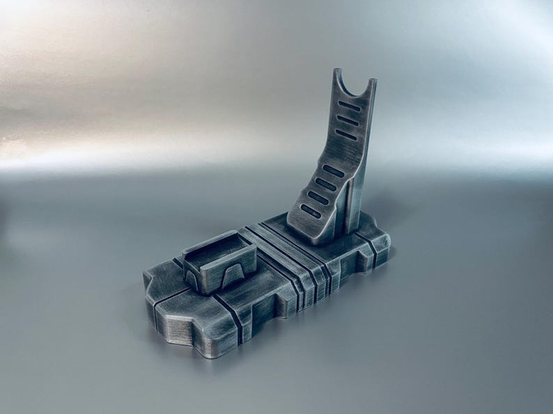 May include: A metallic gray stand with a rectangular base and an angled support. The stand features a small rectangular compartment and a curved, slotted support for holding an item. The design has a futuristic, industrial aesthetic.