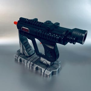 May include: Black Star Wars toy blaster with red accents, resting on a gray stand. The blaster has a detailed design, including a trigger and a sight. The text "STAR WARS" is visible on the side.