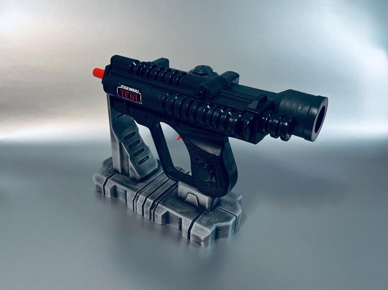 May include: Black Star Wars toy blaster with red accents, resting on a gray stand. The blaster has a detailed design, including a trigger and a sight. The text "STAR WARS" is visible on the side.