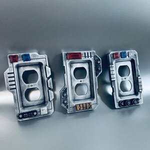 May include: Three decorative electrical outlet covers with a metallic, industrial design. Each cover features a rectangular shape with two circular openings and is accented with red, blue, and gold details. The covers are displayed against a gray background.