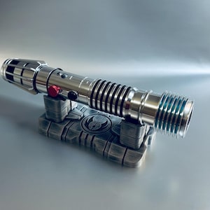 May include: A silver lightsaber with black and red accents rests on a gray stand. The lightsaber features a detailed design with black rings and a red button. The stand has a symbol in the center.