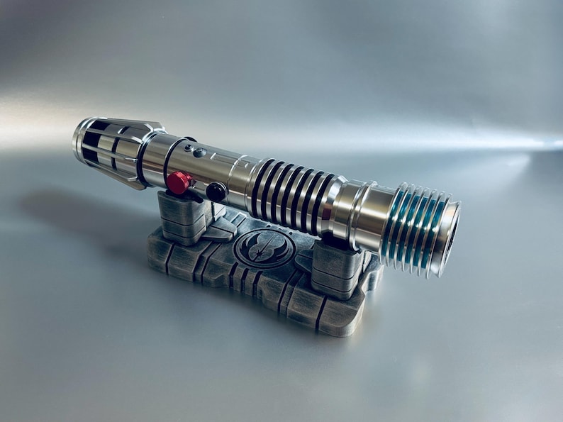 May include: A silver lightsaber with black and red accents rests on a gray stand. The lightsaber features a detailed design with black rings and a red button. The stand has a symbol in the center.