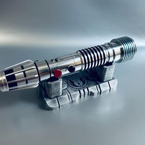 May include: A silver lightsaber with black accents and a red button, resting on a gray stand. The stand features a symbol and is made of a metallic material. The lightsaber has a detailed design.