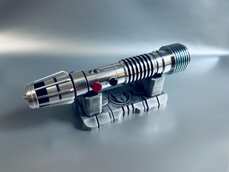 May include: A silver lightsaber with black accents and a red button, resting on a gray stand. The stand features a symbol and is made of a metallic material. The lightsaber has a detailed design.