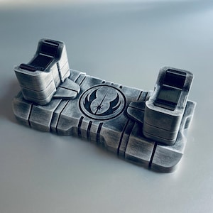 May include: A silver-colored lightsaber display stand with a Star Wars symbol. The stand has two upright holders and a rectangular base with engraved lines. The symbol is a circle with a star and a curved line.