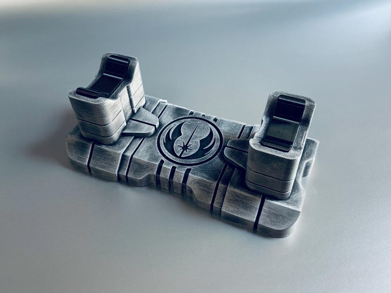 May include: A silver-colored lightsaber display stand with a Star Wars symbol. The stand has two upright holders and a rectangular base with engraved lines. The symbol is a circle with a star and a curved line.