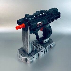 May include: A black Star Wars Jedi blaster toy with a red tip, resting on a gray stand. The blaster has the words "Star Wars Jedi" printed on the side. The stand has a detailed, industrial design.