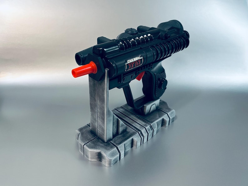May include: A black Star Wars Jedi blaster toy with a red tip, resting on a gray stand. The blaster has the words "Star Wars Jedi" printed on the side. The stand has a detailed, industrial design.