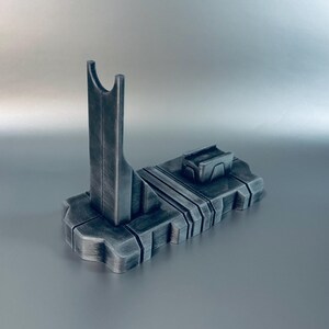 May include: A dark gray, 3D-printed stand with a vertical support and a rectangular base. The stand has a futuristic design with a slot at the top and a small rectangular platform on the base. The stand is made of a textured material.