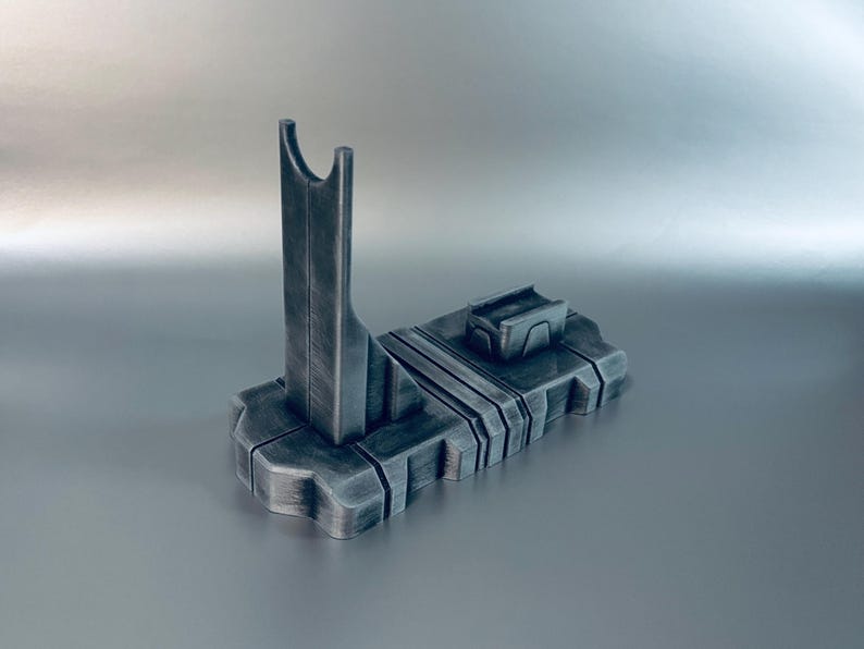 May include: A dark gray, 3D-printed stand with a vertical support and a rectangular base. The stand has a futuristic design with a slot at the top and a small rectangular platform on the base. The stand is made of a textured material.