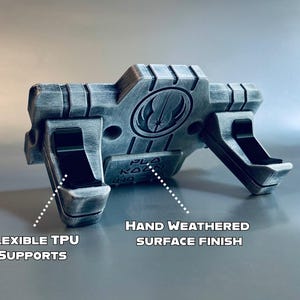 May include: A silver and black display stand featuring a Jedi Order symbol and the text "PLO KOON". The stand includes two black flexible TPU supports and a hand weathered surface finish, designed to hold an object securely in place.