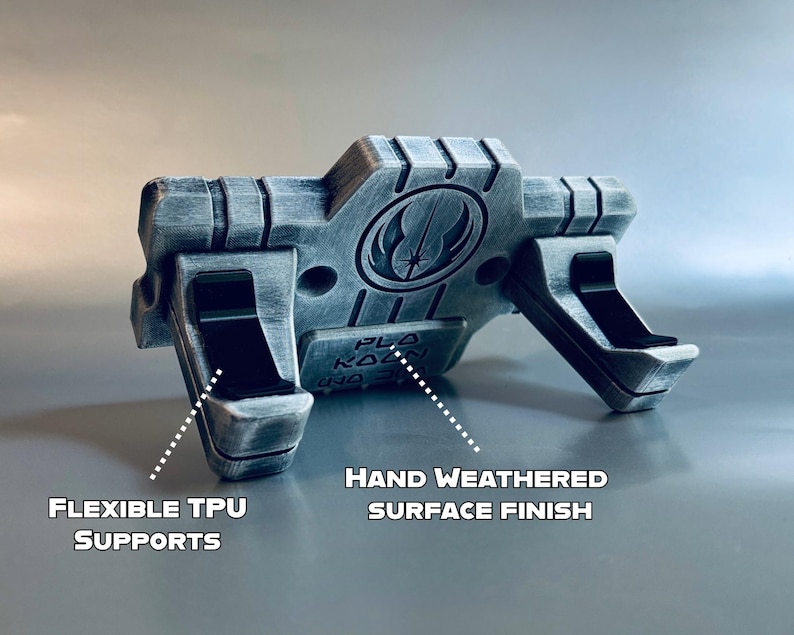 May include: A silver and black display stand featuring a Jedi Order symbol and the text "PLO KOON". The stand includes two black flexible TPU supports and a hand weathered surface finish, designed to hold an object securely in place.