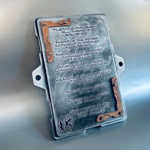 May include: A rectangular, metallic plaque with engraved text and copper-colored corner accents. The text includes philosophical statements. A symbol is visible at the bottom left. The plaque is gray with a brushed metal appearance.