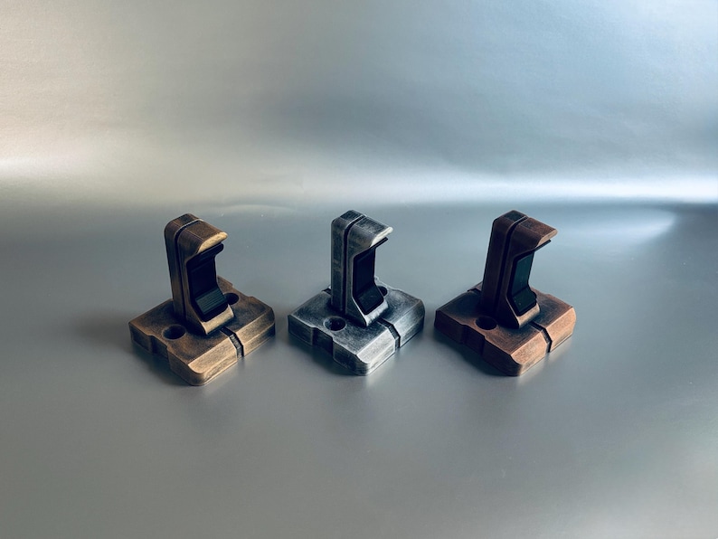 May include: Three metal wall hooks in bronze, silver, and copper tones. Each hook has a square base and a curved top with a black interior. The hooks are designed for hanging items.