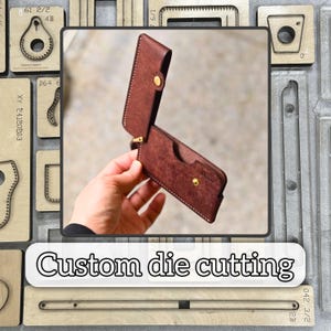 Puede incluir: Una funda de teléfono de cuero marrón con cierre de botón a presión, abierta para revelar el interior. La funda se sostiene en una mano, mostrando las costuras y el diseño. El texto "Custom die cutting" se muestra debajo.