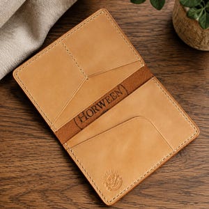 May include: Open, tan leather wallet with stitched edges. The interior features card slots and a central pocket. The word "HORWEEN" is embossed on a brown leather band. A small emblem is visible on the lower right corner.