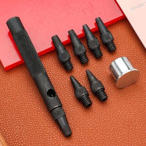 May include: A black metal hole punch tool set with multiple interchangeable tips and a silver thimble. The punch has a textured handle for grip. The set is designed for leathercraft or similar projects.