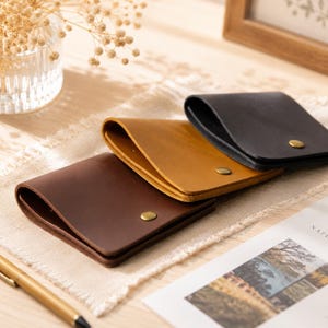 May include: Three leather wallets in brown, tan, and black, each with a curved flap and a brass snap closure. The wallets are arranged on a light-colored surface, with a pen and a decorative floral arrangement in the background.