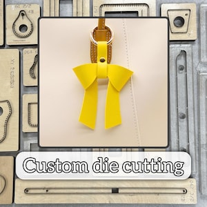 Op de afbeelding: Een gele leren strikvormige sleutelhanger met een goudkleurige ring, bevestigd aan een crèmekleurig leren oppervlak. De tekst "Custom die cutting" wordt weergegeven in een witte doos onderaan de afbeelding. De achtergrond bevat diverse houten stansgereedschappen.