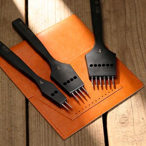 May include: Three black leather working tools with silver prongs are displayed on an orange leather wallet. The tools have the number "4" printed on them. The wallet is on a wooden surface.