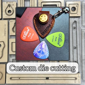 May include: A brown leather guitar pick holder with a gold star emblem and a silver clasp, accompanied by three colorful guitar picks. The picks are orange, green, and blue, with the brand "Alice" printed on them. The text "Custom die cutting" is also visible.